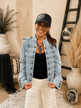 Load image into Gallery viewer, A woman wearing a light-wash blue denim jacket with silver buttons, a black top, white pants, a black cap, and a turquoise necklace, standing in a boho-style room decorated with macrame, dried grass, and vases.