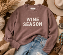 Load image into Gallery viewer, A mauve knit sweater with 'WINE SEASON' in white block letters, styled in a flat lay with light blue jeans, beige boots, a hat, and dried grass on a cowhide rug.