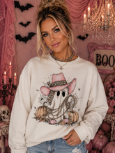 Load image into Gallery viewer, A woman wearing a cream-colored crewneck sweatshirt with a graphic of a ghost in a pink cowboy hat and leopard-print bandana, surrounded by pumpkins and a lasso. She is posing in a pink, Halloween-themed room decorated with bats, a chandelier, and candles.