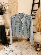 Load image into Gallery viewer, A light blue denim jacket with double-breasted gold buttons, hanging on a wooden hanger in a styled boho room setting with a cowhide rug, woven wall hanging, and dried plants.