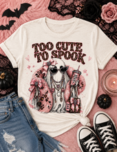 Load image into Gallery viewer, A cream-colored graphic t-shirt featuring a cute ghost character with pink leopard-print faux fur, sunglasses, and the text 'TOO CUTE TO SPOOK'. The shirt is styled in a flat-lay arrangement with blue jeans, black sneakers, pink faux fur, lace, and small pumpkins on a pink background.