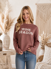 Load image into Gallery viewer, A woman wearing a dusty rose knit sweater with 'WINE SEASON' printed in white across the chest, paired with blue jeans, standing in a cozy, boho-style room decorated with dried pampas grass and a macrame wall hanging.
